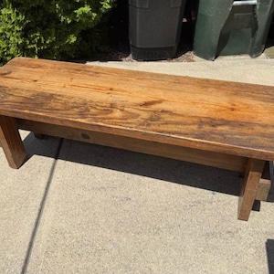 May include: A rectangular wooden bench featuring a natural medium brown wood grain finish. The bench has a smooth, flat top surface with rounded corners and is supported by four sturdy, straight wooden legs positioned at each corner.