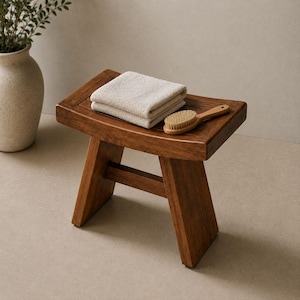 Pode incluir: Um banco de madeira castanho-escuro com um assento retangular de design ripado. Uma toalha bege dobrada e uma pequena escova de banho de madeira com cerdas claras estão colocadas sobre o banco, que tem quatro pernas inclinadas.