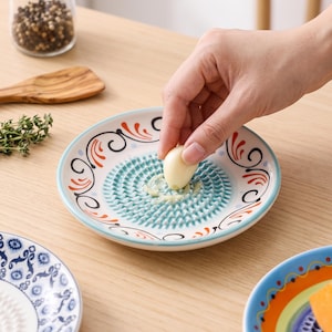 Może przedstawiać: Okrągły ceramiczny talerz do tarcia z teksturowaną powierzchnią pośrodku do ścierania czosnku, otoczony ozdobnym brzegiem z pomarańczowymi i czarnymi wzorami zawijasów na białym tle oraz jasnoniebieskim odcieniem w centrum.