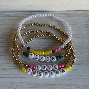 May include: A set of four beaded bracelets featuring white pearl-like beads, metallic gold-tone beads, and colorful seed beads. Two bracelets include white round beads with black lettering that spell out the words TEACH, LOVE, and INSPIRE.