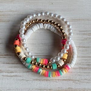 May include: A set of three stacked bracelets featuring a strand of white faux pearls, a strand of white polymer clay discs with gold-tone metallic beads, and a strand of multi-colored clay discs in neon pink, yellow, turquoise, and white.