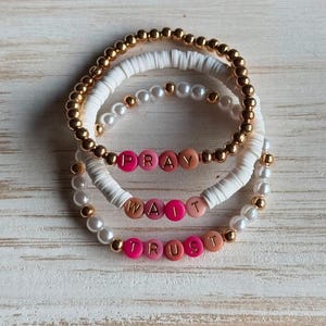 May include: A set of three stacked bracelets featuring gold-tone metallic beads, white disc-shaped polymer clay beads, and white faux pearl beads. Each bracelet includes pink and peach letter beads spelling out the words PRAY, WAIT, and TRUST.