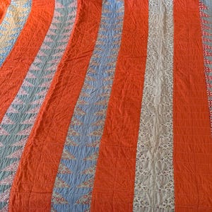 Peut inclure: Une courtepointe composée de bandes verticales alternées en tissu orange vif uni et de bandes à motifs. Les bandes à motifs présentent des formes triangulaires répétitives dans des tons de bleu, blanc et imprimés floraux, assemblées par des coutures en grille.
