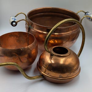 May include: A copper pot with two side handles featuring white and blue floral ceramic accents, a separate copper bowl with a small chain, and a copper watering can with a brass handle and spout, all displayed together on a plain white background.