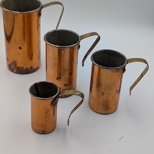 May include: A set of four cylindrical copper measuring cups of varying sizes, each featuring a metallic handle attached to the side. The exterior surfaces show a warm, weathered copper finish with visible signs of patina and minor surface wear.