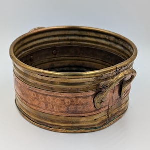 May include: A round, shallow metal basin featuring a central band of hammered copper flanked by smooth brass rims. The vessel includes two curved brass handles attached to the sides, with visible rivets and a weathered, aged patina throughout.
