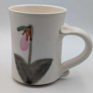 May include: A white ceramic mug featuring a hand-painted illustration of a pink lady slipper orchid with dark green leaves and a brown stem. The mug has a cylindrical body and a curved handle attached to the side, with a smooth, glossy finish.