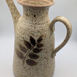 May include: A ceramic jug with a speckled cream and tan glaze, featuring a dark brown leaf imprint on the front. The vessel has a wide, flared rim, a tall spout, and a curved handle, all finished with a consistent speckled texture throughout.