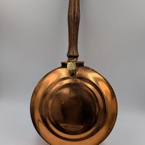 May include: A round copper pan with a polished metallic finish and concentric circular indentations. The pan features a long, vertical wooden handle attached by a brass-coloured metal hinge mechanism at the top edge of the circular body.