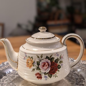 Peut inclure: Une théière en céramique couleur crème avec un corps nervuré et un motif floral composé de roses jaunes, roses et rouge foncé. La théière possède un couvercle assorti avec un bouton doré, une anse courbée et un bec, le tout souligné d'un liseré doré.
