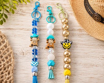 May include: Three beaded keychains with lobster clasps. The left features blue beads, a highland cow charm, and a blue tassel. The center has white and blue beads, a highland cow charm, and a teal tassel. The right has gold beads, a smiley face charm with a cow print hat, and a yellow tassel.
