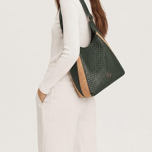 May include: A dark green shoulder bag with a quilted diamond pattern and a single strap. The bag features light brown cork side panels and a small circular logo emblem on the front. The bag is worn over the shoulder with a long strap and buckle.