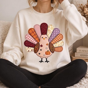 Puede incluir: Una sudadera de cuello redondo color crema con un gráfico central de un pavo con una cola estilo patchwork. El cuerpo del pavo es de color rosa claro con pequeñas ilustraciones de hojas, estrellas y calabazas. Las plumas de la cola tienen varios patrones en naranja, granate, morado y crema.