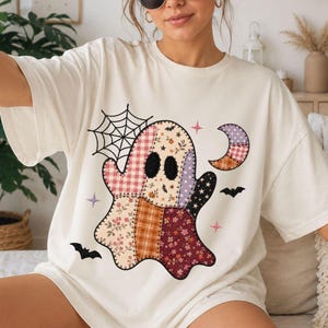 Puede incluir: Una camiseta color hueso con un gran gráfico central de un fantasma compuesto por varios patrones de patchwork, incluyendo cuadros, flores y estrellas. El diseño incluye una telaraña negra, una luna creciente y dos pequeños murciélagos negros.