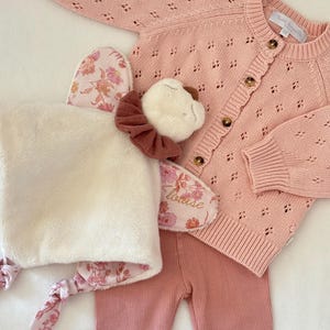 Peut inclure: Un gilet en tricot rose clair avec un motif en losanges et des boutons marron, accompagné d'un legging côtelé assorti et d'un doudou en peluche avec une tête crème, un col à volants marron et un tissu floral rose portant le nom Louise.