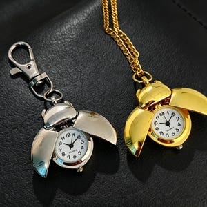May include: A silver-toned beetle-shaped keychain watch and a gold-toned beetle-shaped pendant watch on a chain. Both timepieces feature white dials with black Arabic numerals, black hands, and the word Quartz printed on the face.