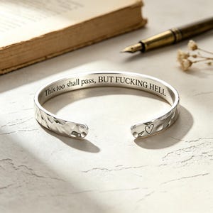 Pode incluir: Uma pulseira de punho prateada com uma textura exterior martelada. O interior está gravado com o texto "This too shall pass, BUT FUCKING HELL" numa fonte serifada. Uma extremidade da pulseira apresenta um pequeno símbolo de coração.