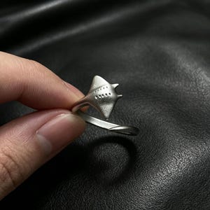 Può includere: Un anello aperto color argento con un design a forma di manta sulla parte superiore. La manta presenta una finitura opaca con piccoli dettagli circolari incavati sul dorso. Il gambo dell'anello è liscio e si avvolge a formare la creatura marina.