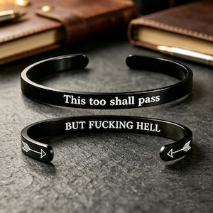 Könnte beinhalten: Zwei schwarze Armreifen aus Metall. Der obere Armreif ist mit dem weißen Text "This too shall pass" graviert. Der untere Armreif ist mit dem weißen Text "BUT FUCKING HELL" graviert und hat an beiden offenen Enden ein weißes Pfeilsymbol.