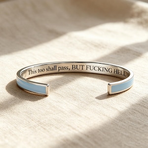 May include: A silver-toned cuff bracelet featuring a light blue enamel inlay on the exterior and the engraved black text "This too shall pass, BUT FUCKING HELL" on the interior surface. The open-ended bangle has a smooth, polished finish.