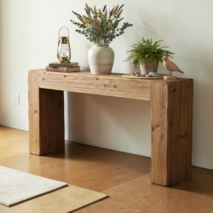 May include: A rectangular console table constructed from thick, light brown reclaimed wood with a visible grain and textured surface. The table features two sturdy, block-style legs supporting a long, solid top, providing a stable base for decor.