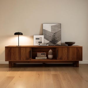 May include: A long wooden media console with vertical slatted sliding doors, featuring a black mushroom-shaped lamp, two framed geometric prints, a small radio, a two-toned ceramic vase, and a dark bowl arranged on its flat top surface.