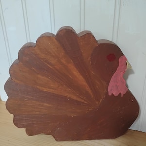 Op de afbeelding: Een platte, uitgesneden houten kalkoendecoratie geschilderd in bruintinten. De kalkoen heeft een waaiervormige staart, een rode vlek bij het oog, een lichtroze lellen en een kleine, puntige snavel. Het oppervlak toont zichtbare penseelstreken.