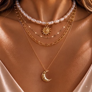 May include: A layered necklace set featuring a top strand of white oval pearls with a gold sun pendant, followed by a thin gold chain, a gold paperclip link chain, and a bottom gold chain with a textured crescent moon pendant.