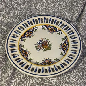 May include: A round ceramic plate with a white center featuring a central floral basket motif and colorful decorative borders. The rim is designed with a series of vertical cutouts accented by blue paint, creating a lattice-like appearance.