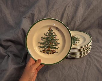 Puede incluir: Una pila de platos de cerámica de color crema con un borde verde y una ilustración central de un árbol de Navidad decorado con adornos coloridos y regalos envueltos en la base. Los platos tienen un borde liso y un acabado brillante.