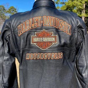 Könnte beinhalten: Eine schwarze Lederjacke mit dem Harley-Davidson-Logo, das in Orange und Weiß auf den Rücken gestickt ist. Der Schriftzug Harley-Davidson ist über dem zentralen Schildemblem gewölbt, darunter ist das Wort Motorcycles in Orange gestickt.