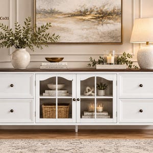 May include: A white wooden media console featuring a dark brown top, four drawers with small black knobs, and two central glass-paneled cabinet doors. The interior shelves display books, a woven basket, and decorative items, supported by turned feet.