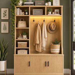 May include: A light oak entryway hall tree featuring a tall storage unit with open shelving, three black coat hooks, and a lower cabinet with three fluted doors. The unit includes integrated lighting, a beige handbag, a hat, and a woven basket.