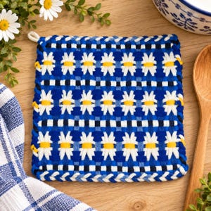 May include: A square woven potholder featuring a repeating pattern of white daisy flowers with yellow centers against a deep blue background. The textile has a thick, braided border in alternating shades of blue and white, creating a textured edge.