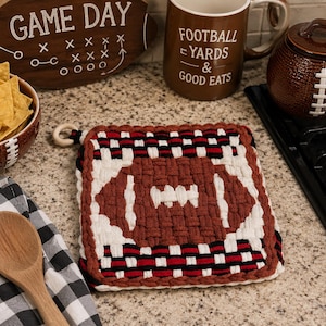 May include: A square woven potholder featuring a brown football design with white laces in the center. The border consists of alternating rows of black, white, and red fabric strips. A small wooden ring is attached to the top left corner for hanging.