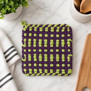 May include: A square potholder featuring a woven grid pattern in deep purple and bright lime green. The potholder has a thick, textured construction with a small loop at the top corner for hanging, showcasing a consistent checkerboard design.