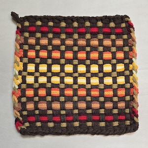 May include: A square potholder featuring a woven grid pattern in dark brown, tan, red, orange, and yellow. The design consists of horizontal and vertical interlaced strips creating a checkerboard effect with a braided border edge on all sides.