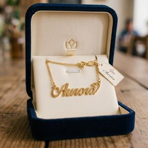 May include: A gold-toned name necklace featuring the script name Aurora with a small heart detail at the end. The necklace is displayed inside a navy blue velvet jewellery box with a cream-coloured interior and a tag labelled For Aurora.
