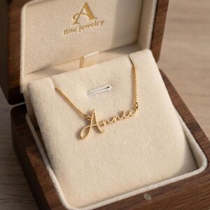 May include: A gold-toned name necklace featuring the name Annie in a cursive script font, attached to a delicate box chain. The necklace is displayed inside a dark wooden jewellery box with a cream-coloured velvet interior and the text A fine jewellery.