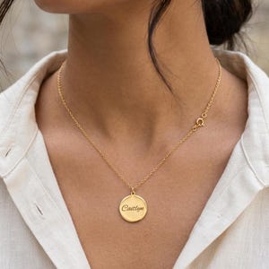 May include: A gold-toned necklace featuring a circular pendant with the name Caitlyn engraved in a cursive script. The pendant is suspended from a delicate, thin gold-toned chain that secures with a small round spring ring clasp at the back.