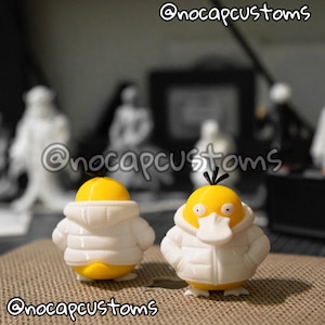 May include: Two yellow duck-like figures wearing white puffer jackets with hoods. The figures have small black tufts on their heads and white beaks. One figure is shown from the back, and the other is shown from the front. Text reads @nocapcustoms.