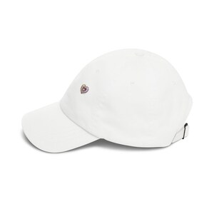 May include: A white baseball cap featuring a small, embroidered heart design on the front panel. The heart is outlined in gold with a purple center and small radiating lines. The cap has a curved brim and an adjustable strap at the back.