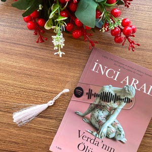 May include: A clear acrylic bookmark featuring a black Spotify code and icon, attached to a white tassel. The bookmark is placed on the cover of the book titled Inci Aral Verda'nın Ölümü, which displays a pink background and a floral figure.