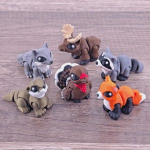 May include: A collection of six articulated animal figurines including a gray wolf, a brown moose, a black and white raccoon, a brown turkey with a red wattle, a tan otter, and an orange fox, all featuring large black eyes and segmented bodies.
