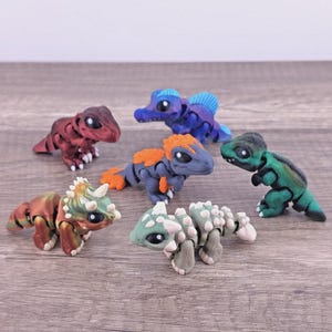 May include: A collection of five articulated dinosaur figurines featuring a red T-Rex, a blue Spinosaurus, a grey and orange raptor, a green T-Rex, and a multi-colored triceratops, all with large black eyes and detailed textured skin patterns.