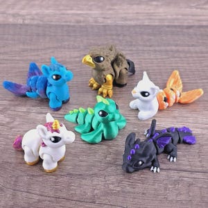 May include: A collection of six articulated plastic creature figurines including a blue aquatic animal, a brown eagle-like bird, a white and orange fish-like creature, a green dragon, a white unicorn with a gold horn, and a black dragon with purple accents.