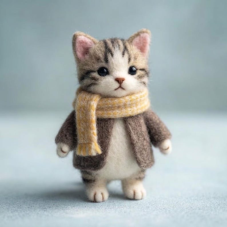 以下が含まれることがあります： 茶色と白の縞模様の毛並みを持つニードルフェルトの子猫のフィギュア。質感のある茶色のジャケットと、黄色と白のストライプ柄のマフラーを着用している。暗い色の目、小さなピンクの鼻、尖った耳をしており、直立した姿勢で立っている。