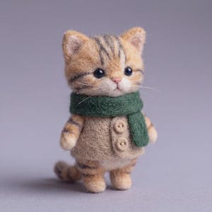 以下が含まれることがあります： 茶色と黄褐色の縞模様を持つ小さなトラ猫のニードルフェルト人形。濃い緑色のフェルト製スカーフと、前面に2つの小さな丸いボタンが付いたベージュのフェルト製ベストを着用している。猫には暗い色の目と白いひげがある。