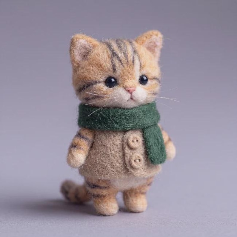 以下が含まれることがあります： 茶色と黄褐色の縞模様を持つ小さなトラ猫のニードルフェルト人形。濃い緑色のフェルト製スカーフと、前面に2つの小さな丸いボタンが付いたベージュのフェルト製ベストを着用している。猫には暗い色の目と白いひげがある。