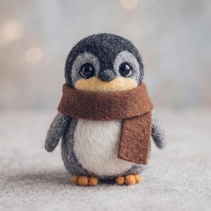 Puede incluir: Una figura de pingüino de lana fieltrada con cuerpo gris oscuro y blanco, pico amarillo suave y dos ojos negros. El pingüino lleva una bufanda de fieltro marrón sólido envuelta alrededor del cuello y tiene pequeños pies naranjas en la base.